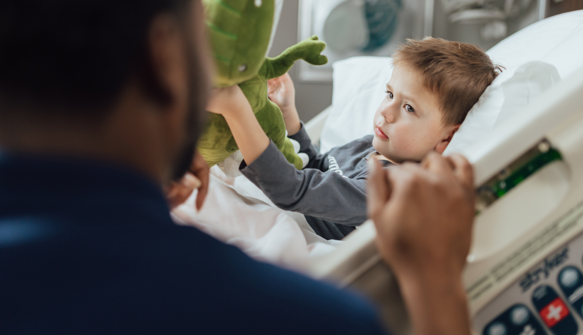 Building a Culture of Safety Hero Image Adorable toddler boy curiously reaches for stuffed green dinosaur toy held by unidentified person.
