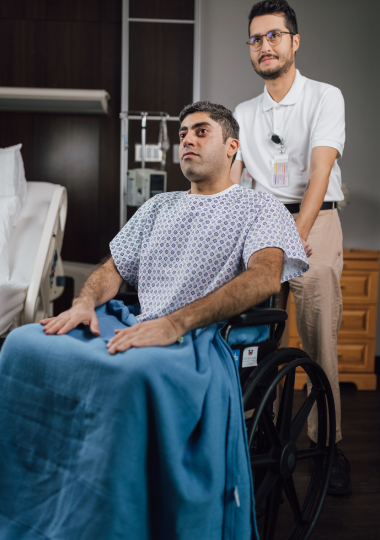 Middle-aged patient in hospital gown sits in wheelchair while male nurse stands behind him