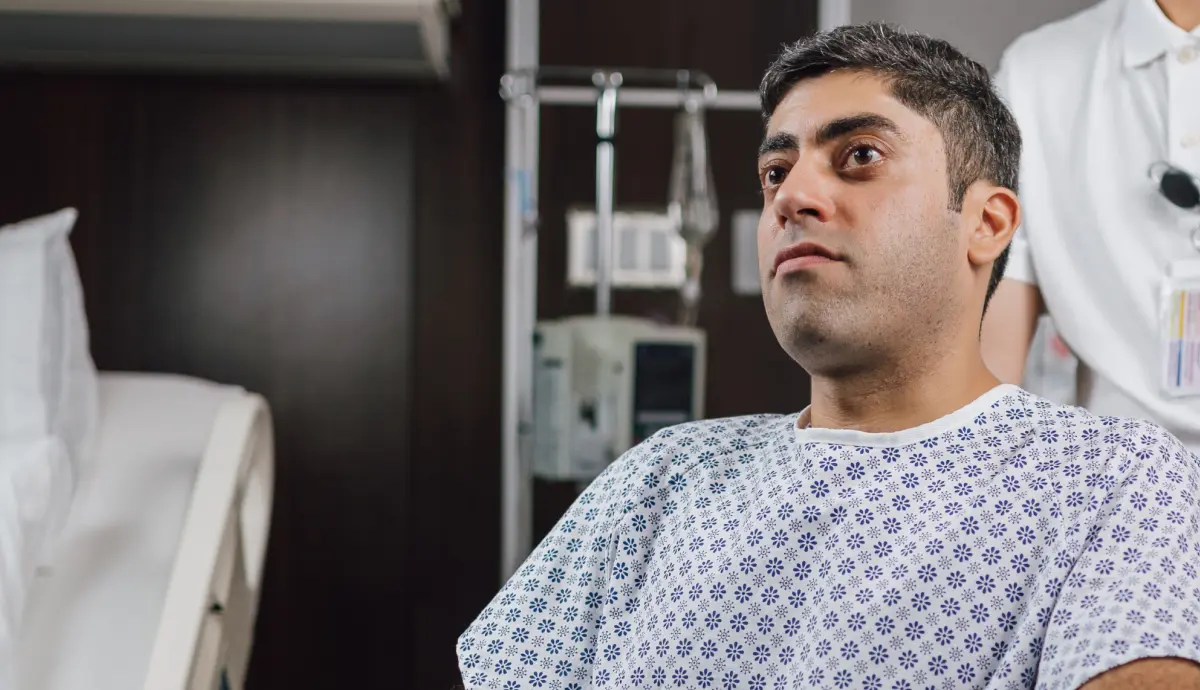 Pensive male patient in hospital gown sitting on bed in dimly lit hospital room