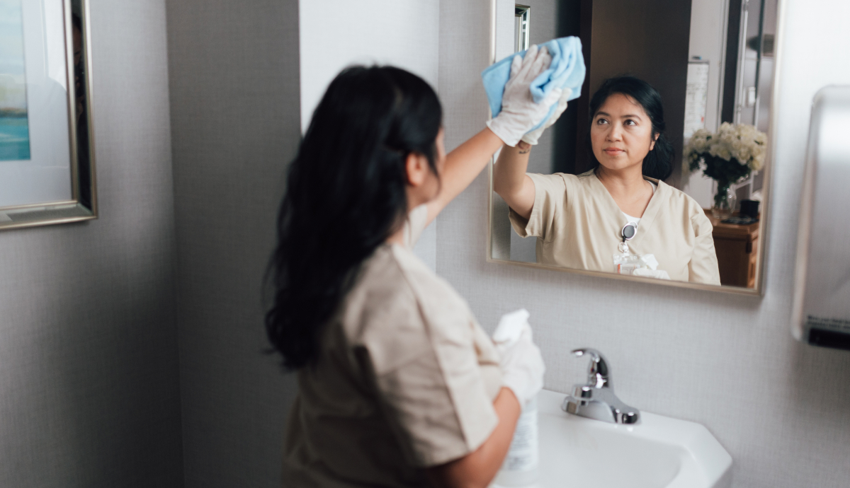 Young female healthcare worker cleaning mirror while looking at reflection in medical facility bathroom