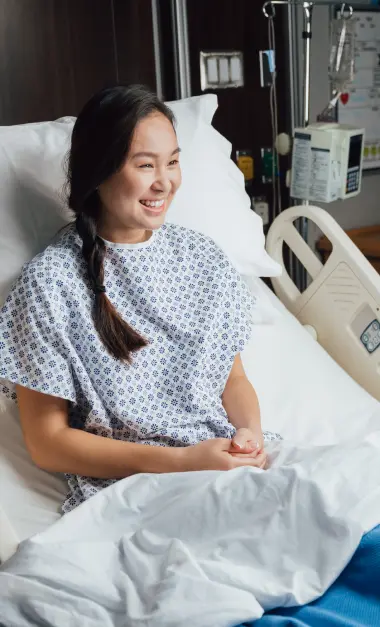 Smiling Asian patient in hospital gown sitting up in bed, attached to IV drip