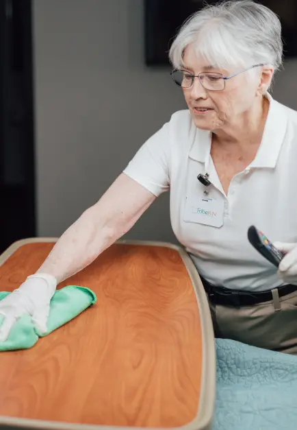 Alt text: Elderly woman in Crothall Healthcare uniform cleaning table with green cloth in healthcare facility