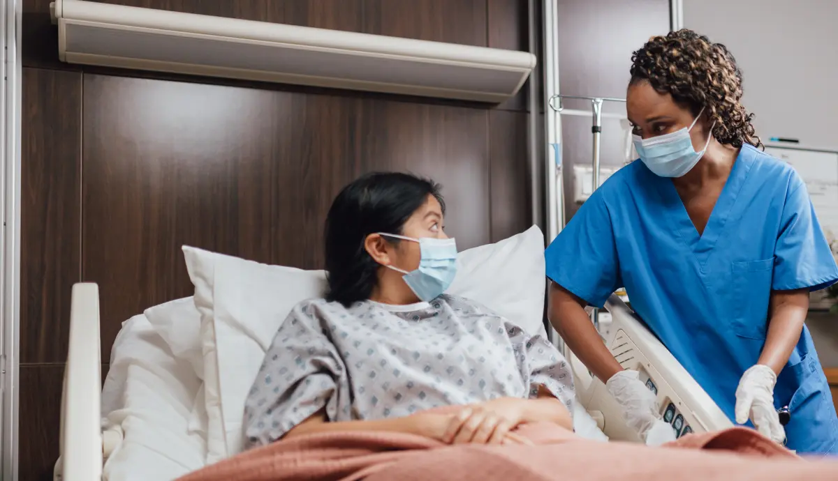 Practice Makes Perfect Hero Image Masked nurse checks on patient in hospital bed while wearing blue scrubs and gloves