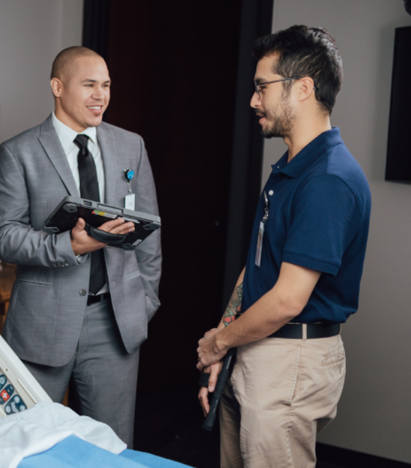 Two men chatting, one in a suit holding a tablet and the other casually dressed, in an office setting.