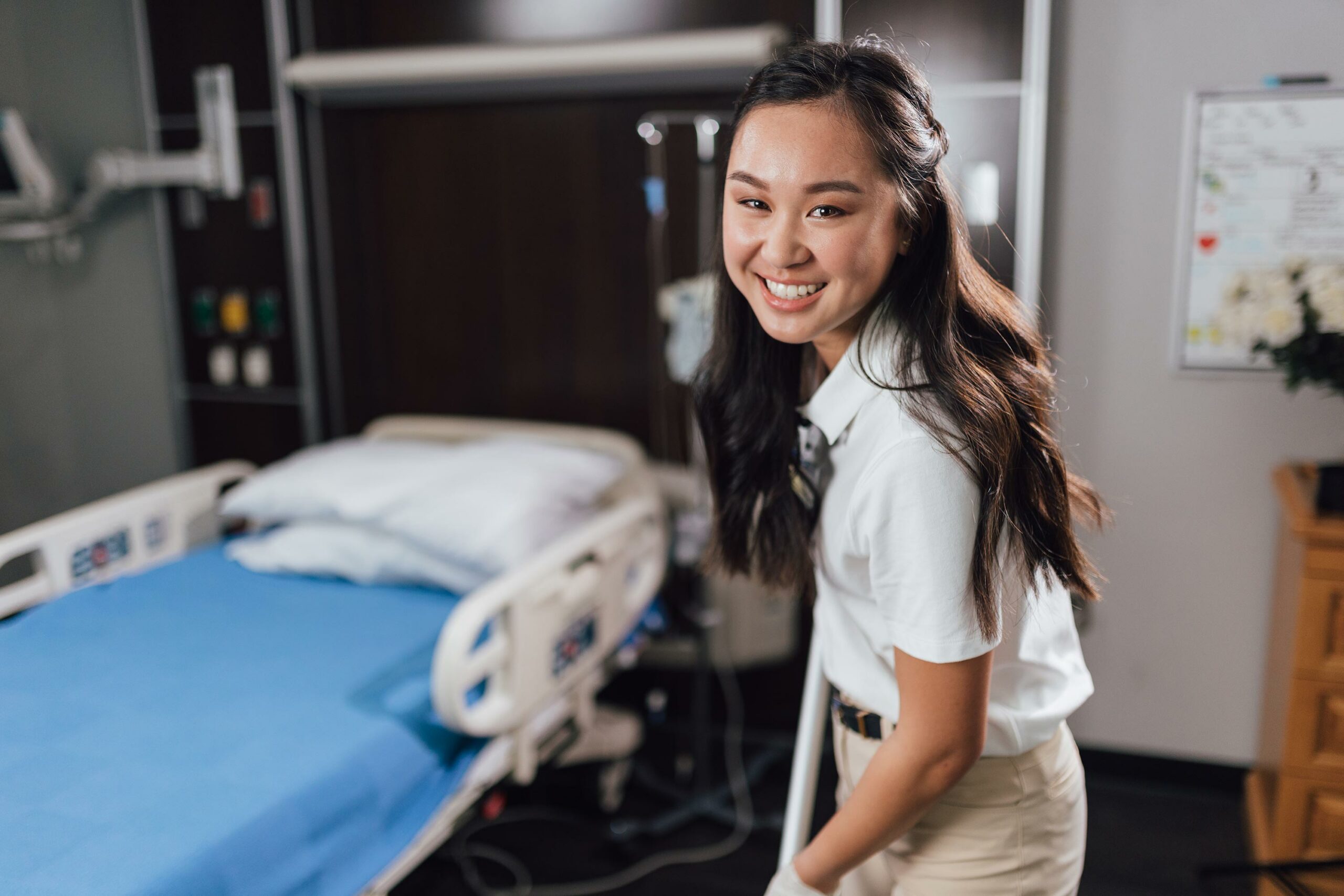 Smiling young Asian female healthcare worker standing next to hospital bed, providing friendly patient care.
