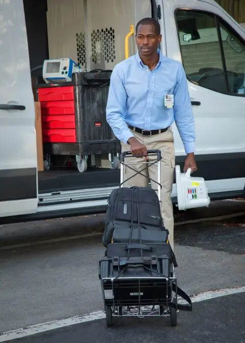 Hospital employee standing with equipment cart and luggage, preparing medical transport vehicle.