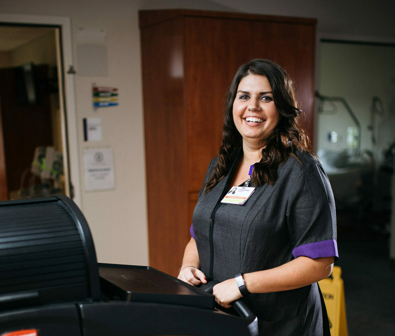 IP1A4273 Smiling healthcare worker holding clipboard at nurse's station, wearing ID badge