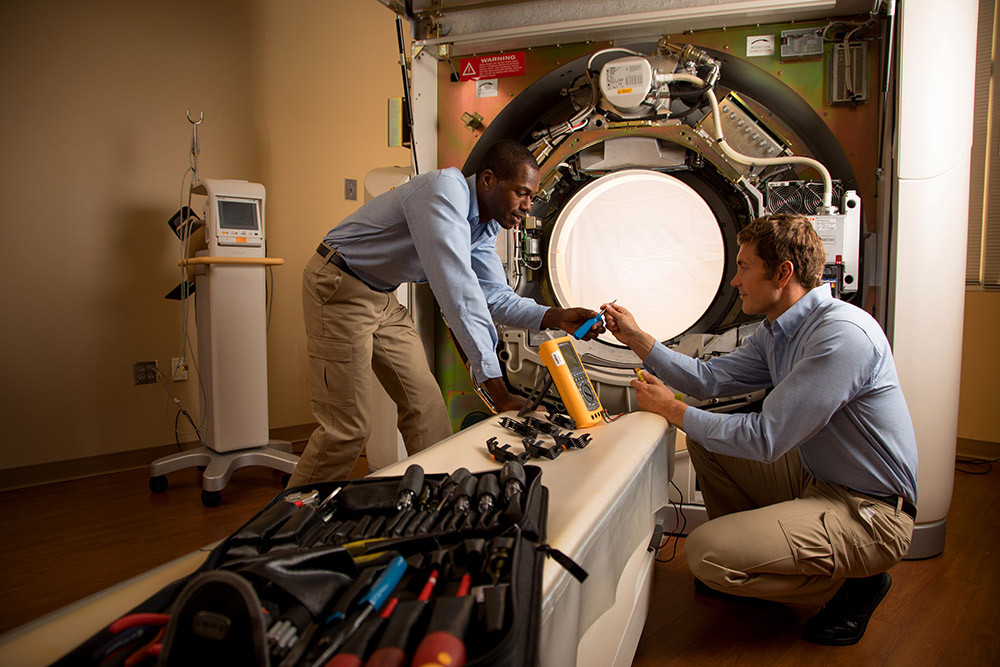 Two technicians performing maintenance on medical imaging equipment in a hospital room.