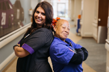 Alt text: Smiling healthcare worker assists elderly patient in hospital hallway